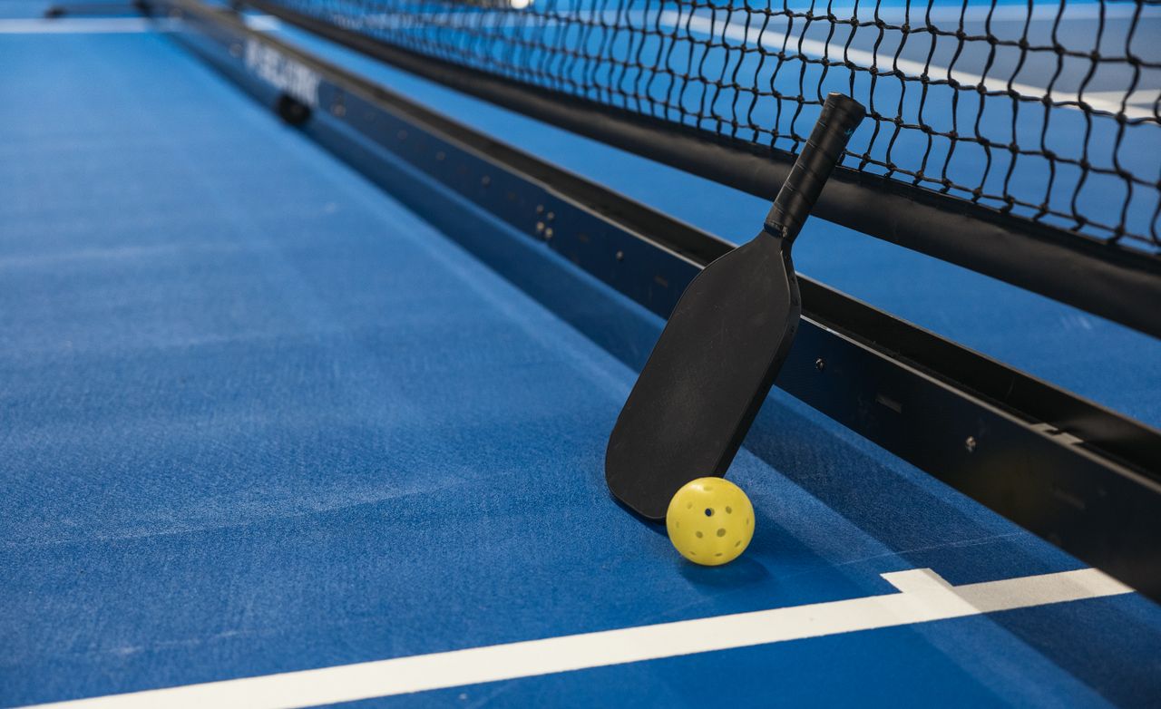 heros-img Pickleball paddle resting against net with yellow ball on blue court