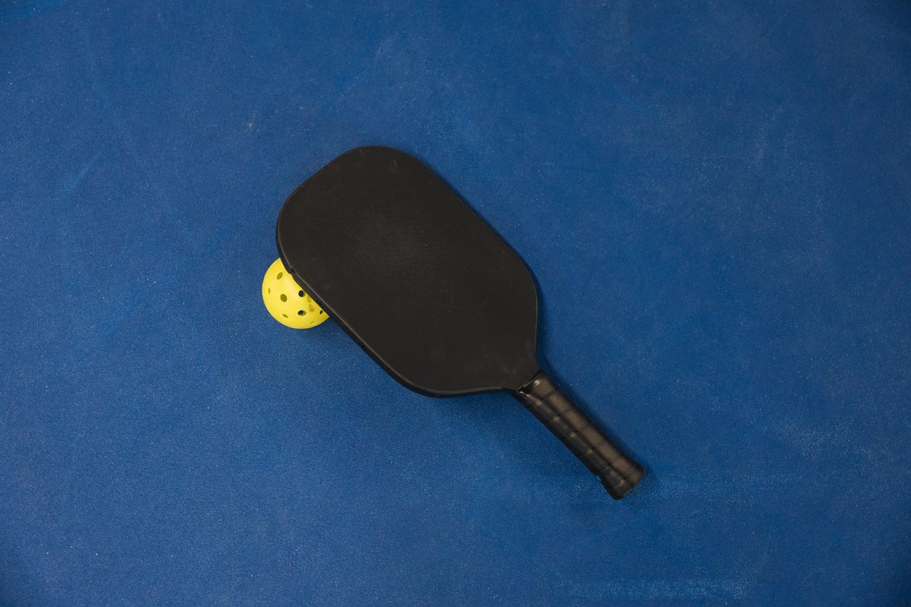 about-us-01 A pickleball paddle and ball on a textured blue surface, showcasing sports equipment.