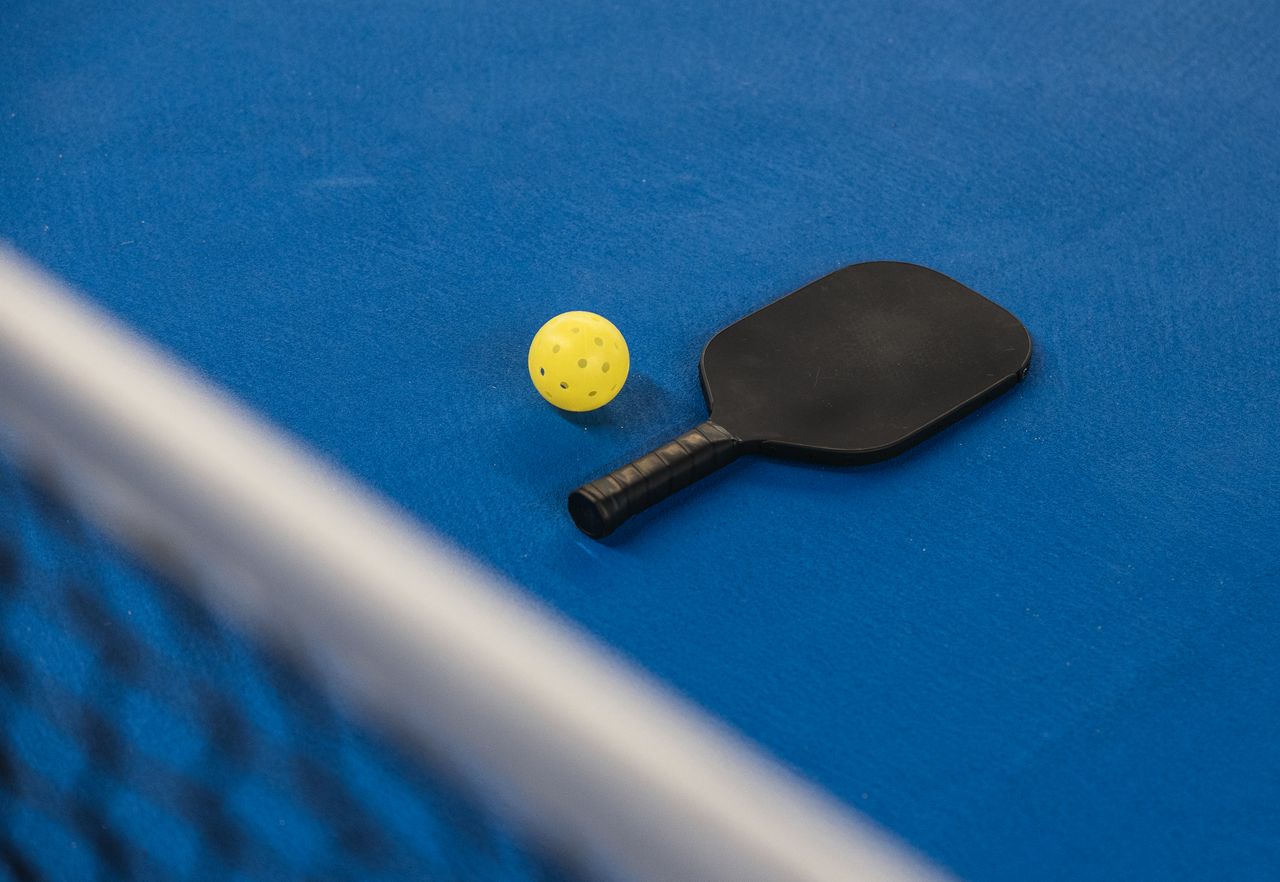 services-01 A pickleball paddle and ball resting on a blue court surface with a net in focus.