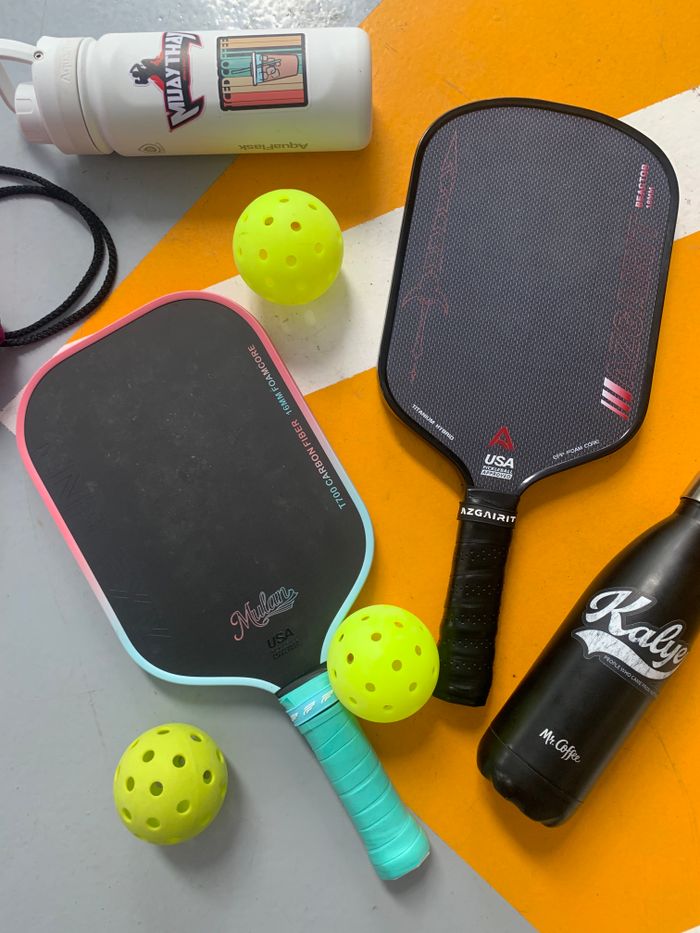 about-us-02 Pickleball paddles and balls arranged with water bottles on a colorful floor.